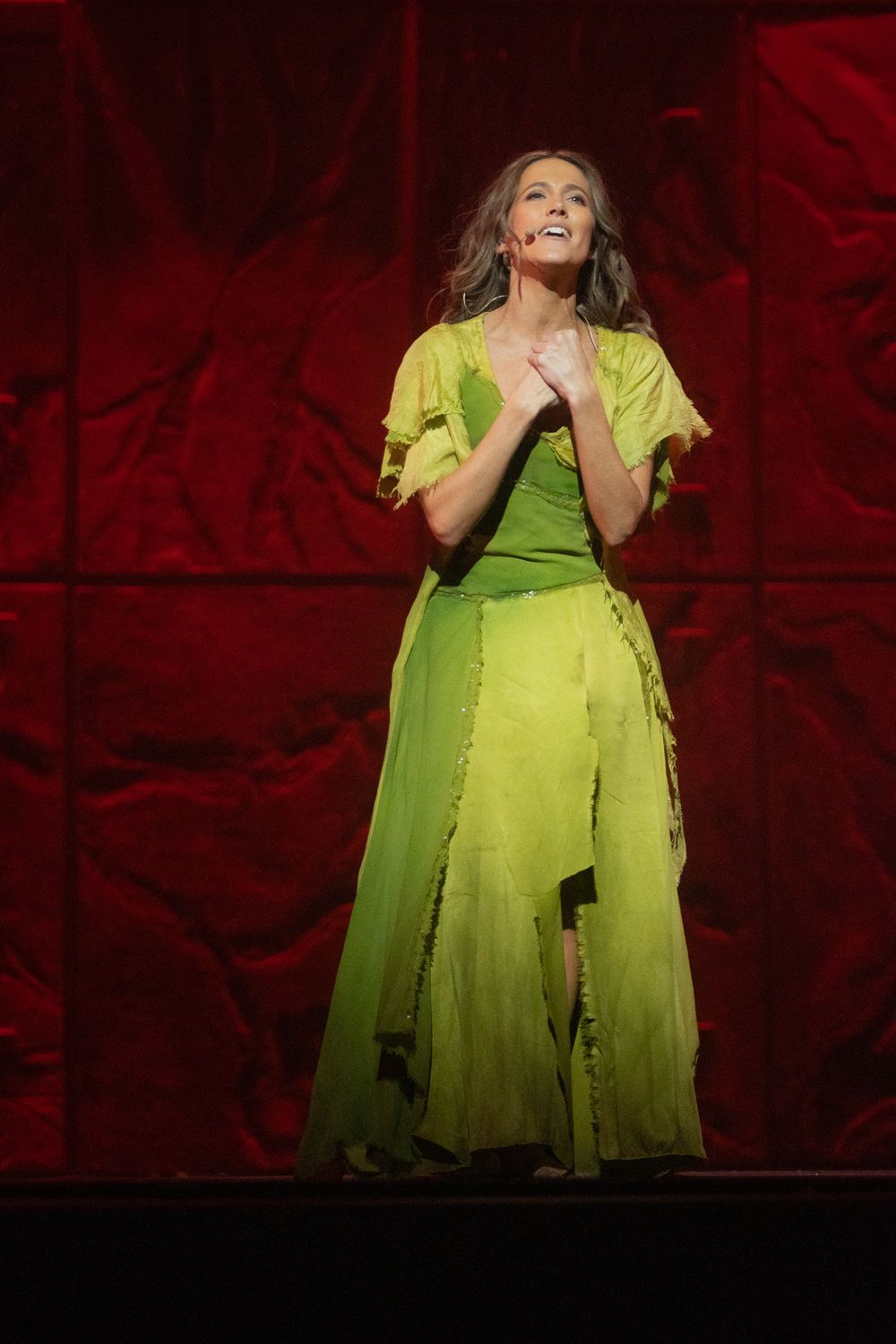 Chanteuse aux longs cheveux ondulés, micro-serre-tête, robe verte déchirée, mains jointes sur scène devant un décor rouge
