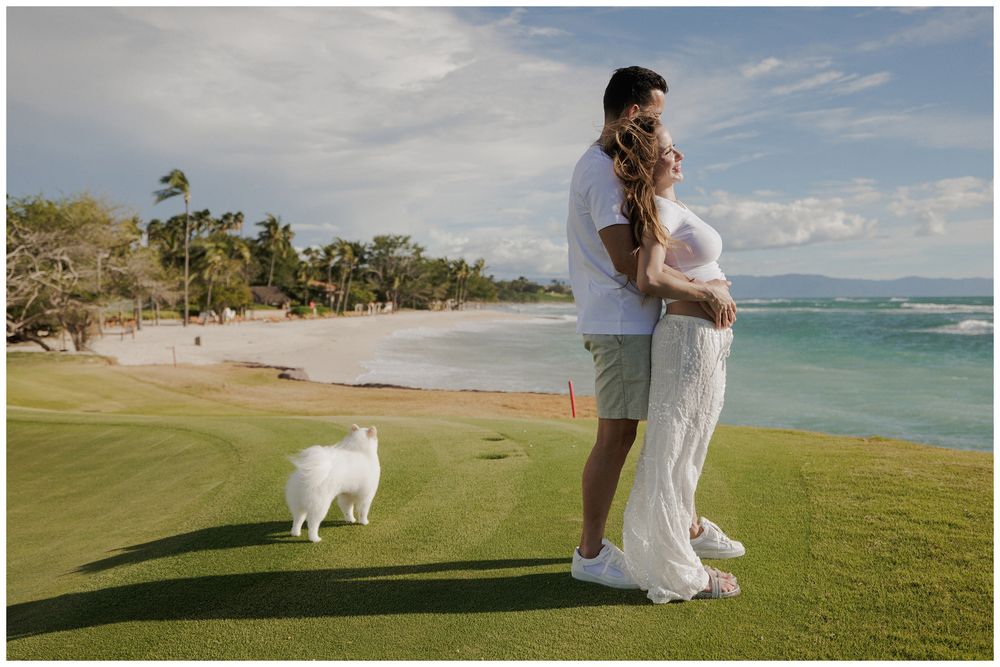 A maternity photoshoot at Punta Mita, featuring a couple embracing on a tropical lawn with their fluffy white dog near the beach.