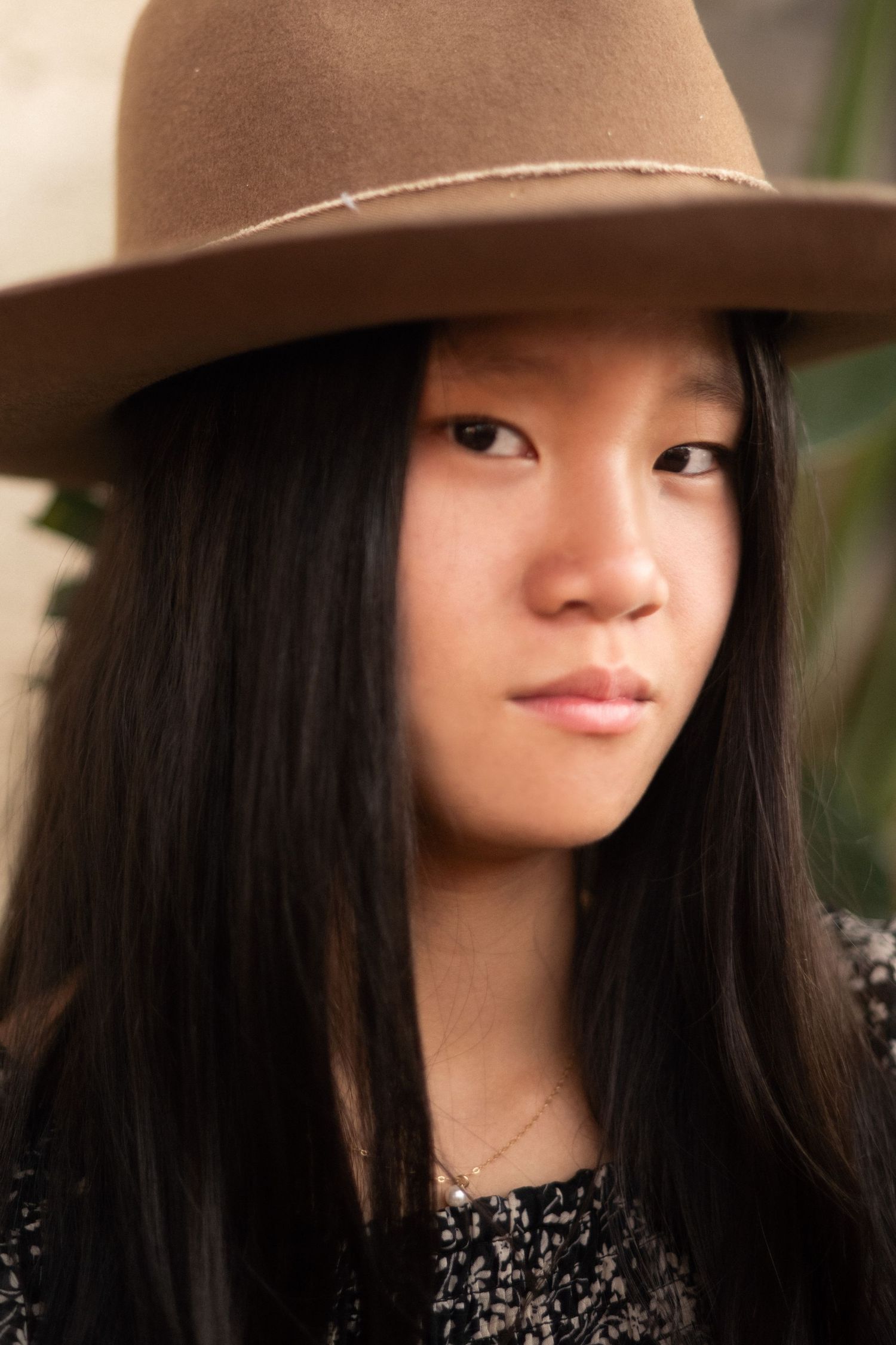 Young Asian woman with long black hair wearing a tan wide-brim felt hat, looking directly at camera.