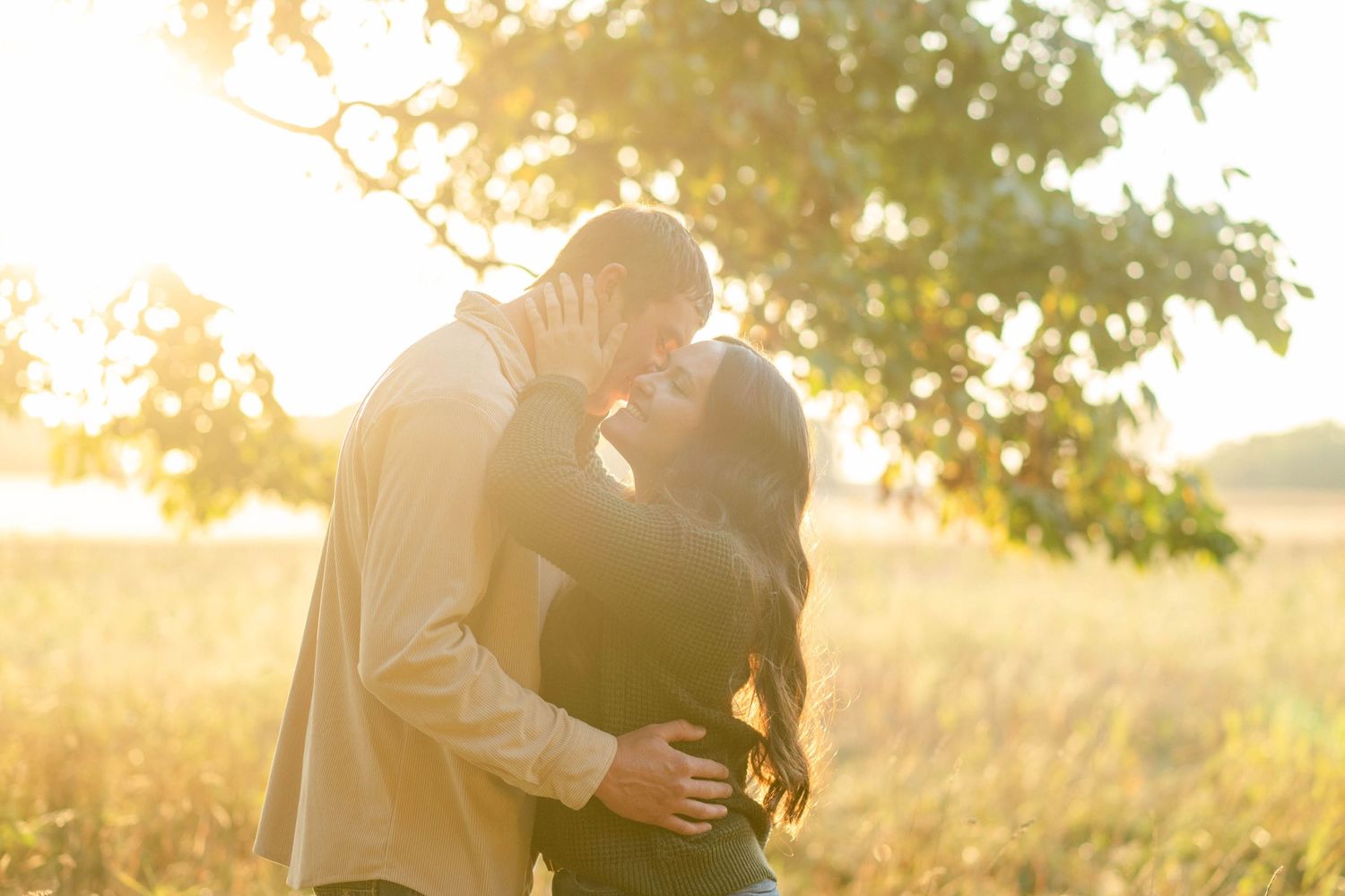 Couple shares a kiss backlit by glowing sunset under a tree during outdoor engagement session.