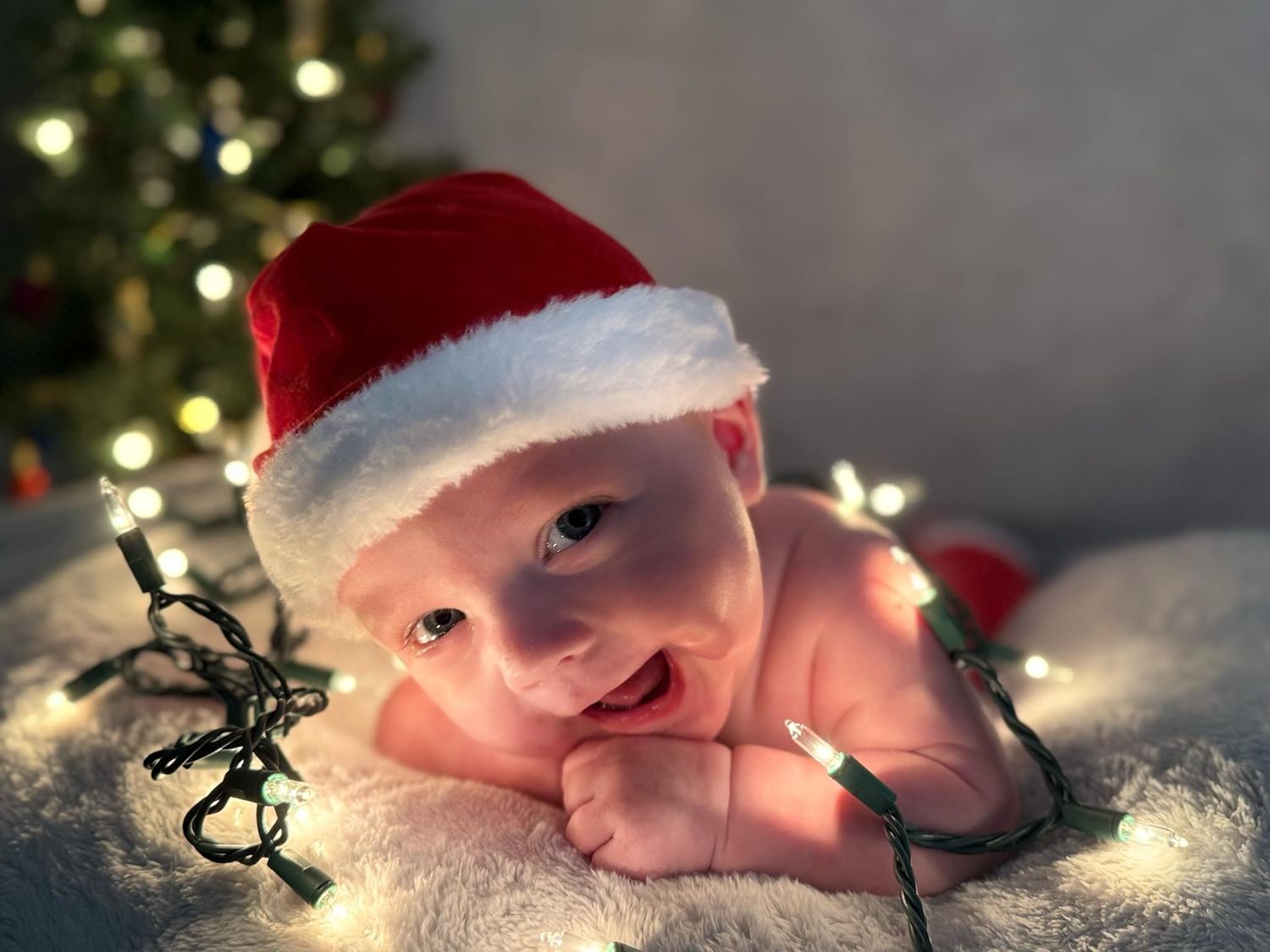 Smiling baby in a red Santa hat lies on a soft blanket wrapped in string lights, with a glowing Christmas tree in the background.