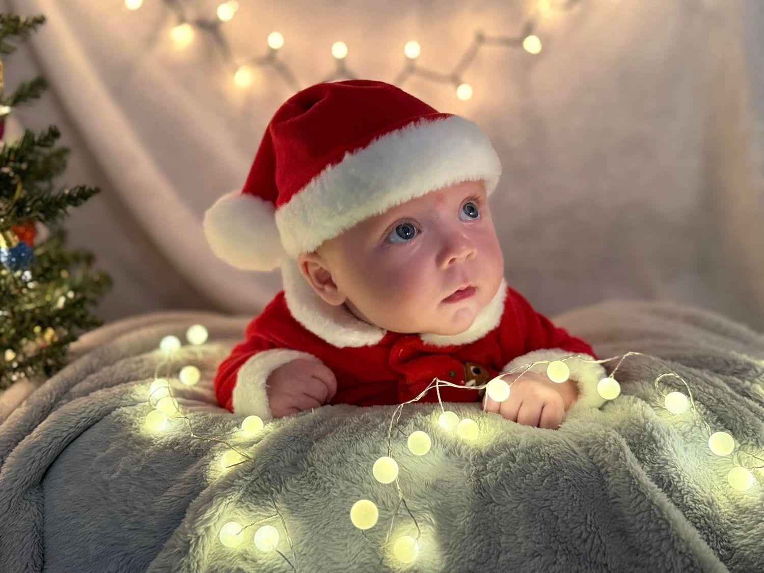 Baby in a red Santa suit and hat lies on a gray blanket surrounded by glowing fairy lights beside a small Christmas tree.