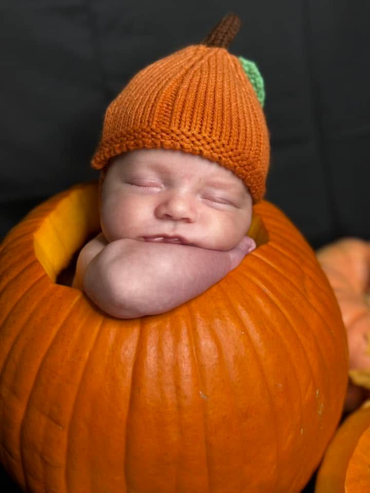 Sleeping newborn baby in a knit orange pumpkin hat rests inside a hollowed-out pumpkin for an autumn-themed photo.