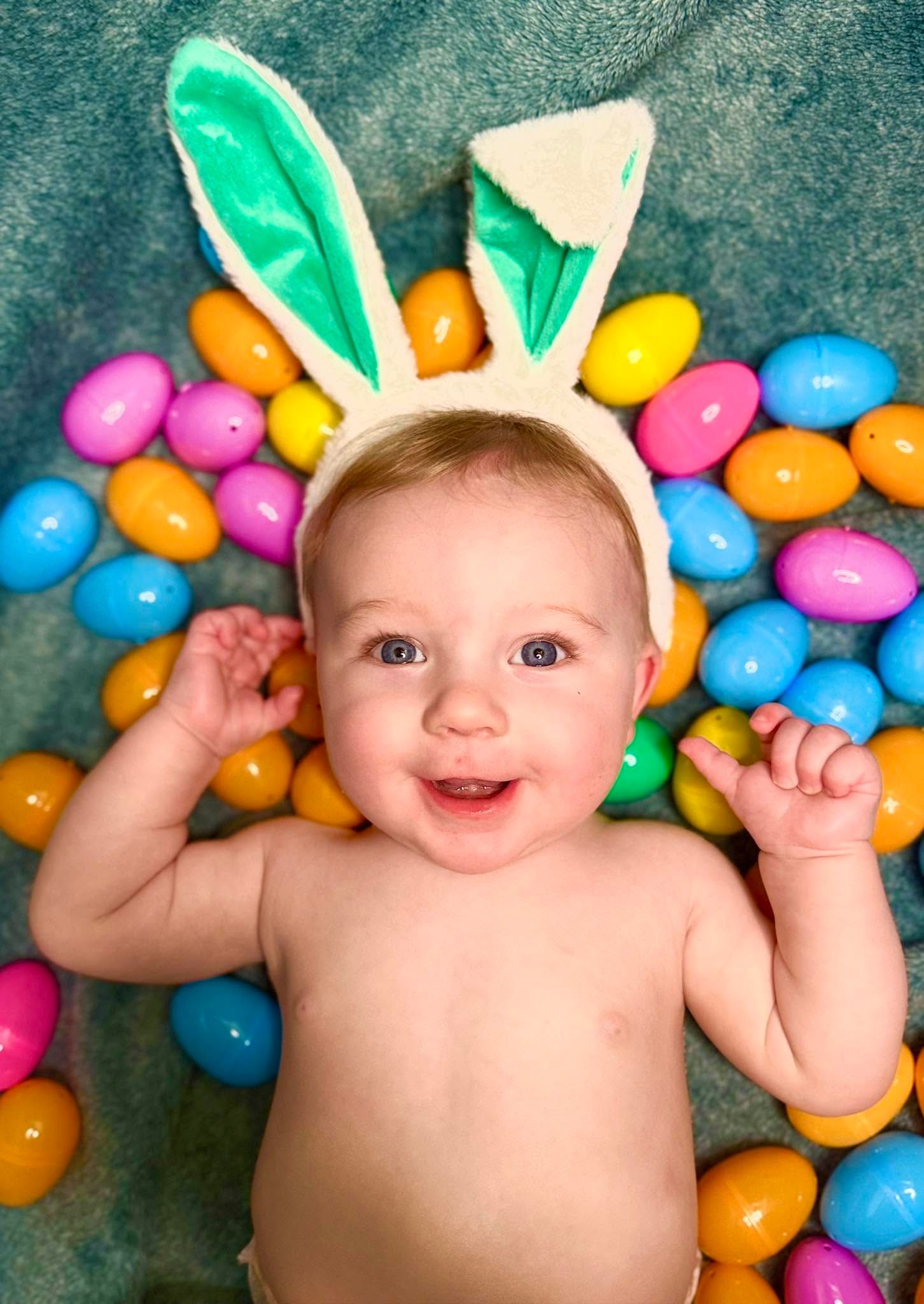Smiling baby wearing white bunny ears lies on a teal blanket surrounded by colorful plastic Easter eggs in a festive photo.