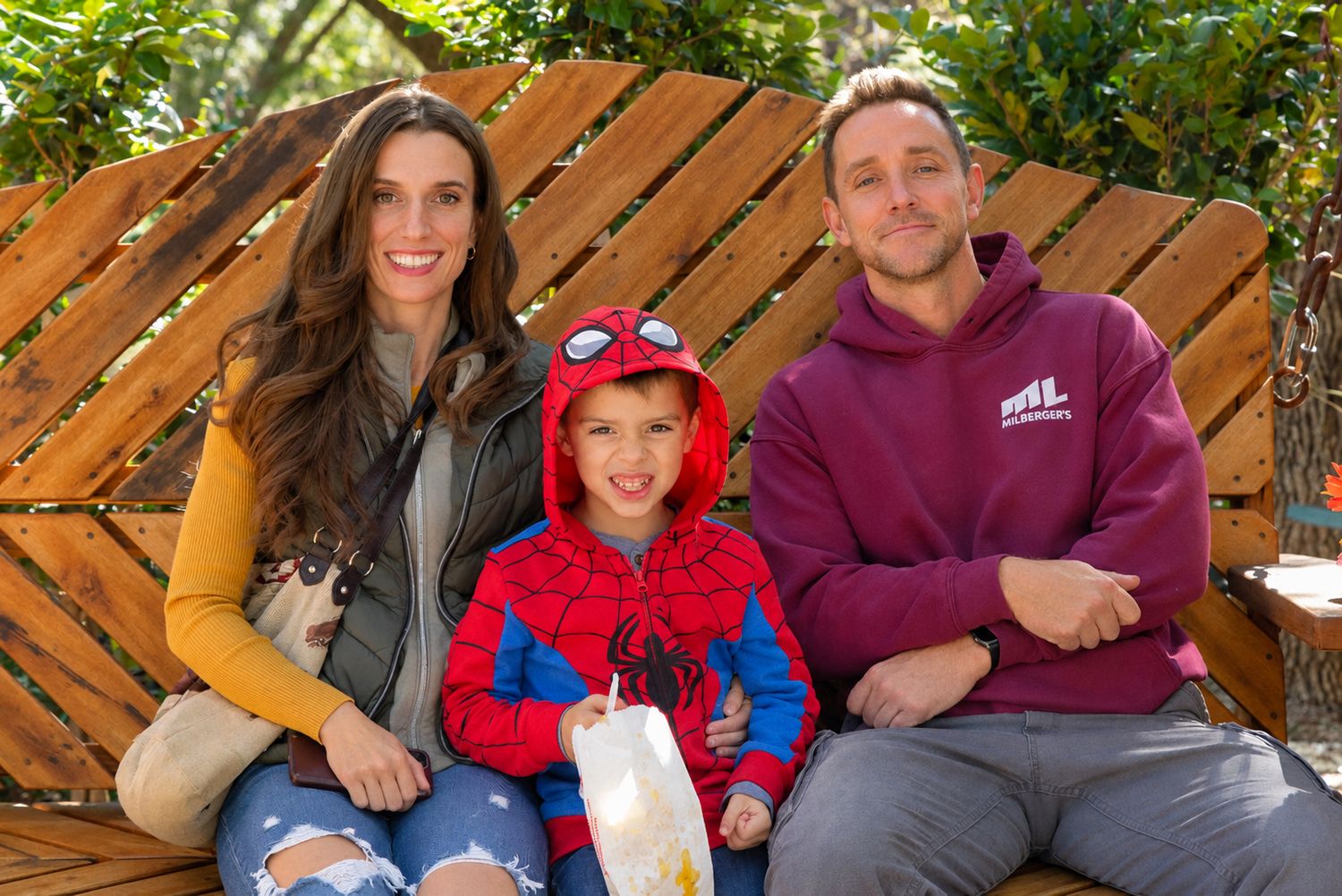 A smiling woman and man sit on a wooden bench with a young boy in a Spider-Man hoodie holding a snack bag outdoors.