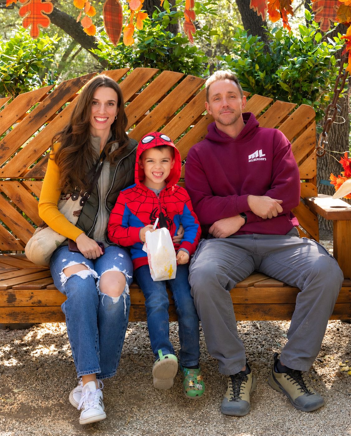 A smiling family of three sits on a wooden bench outdoors, with a young boy in a red hooded jacket holding a white bag between his parents.