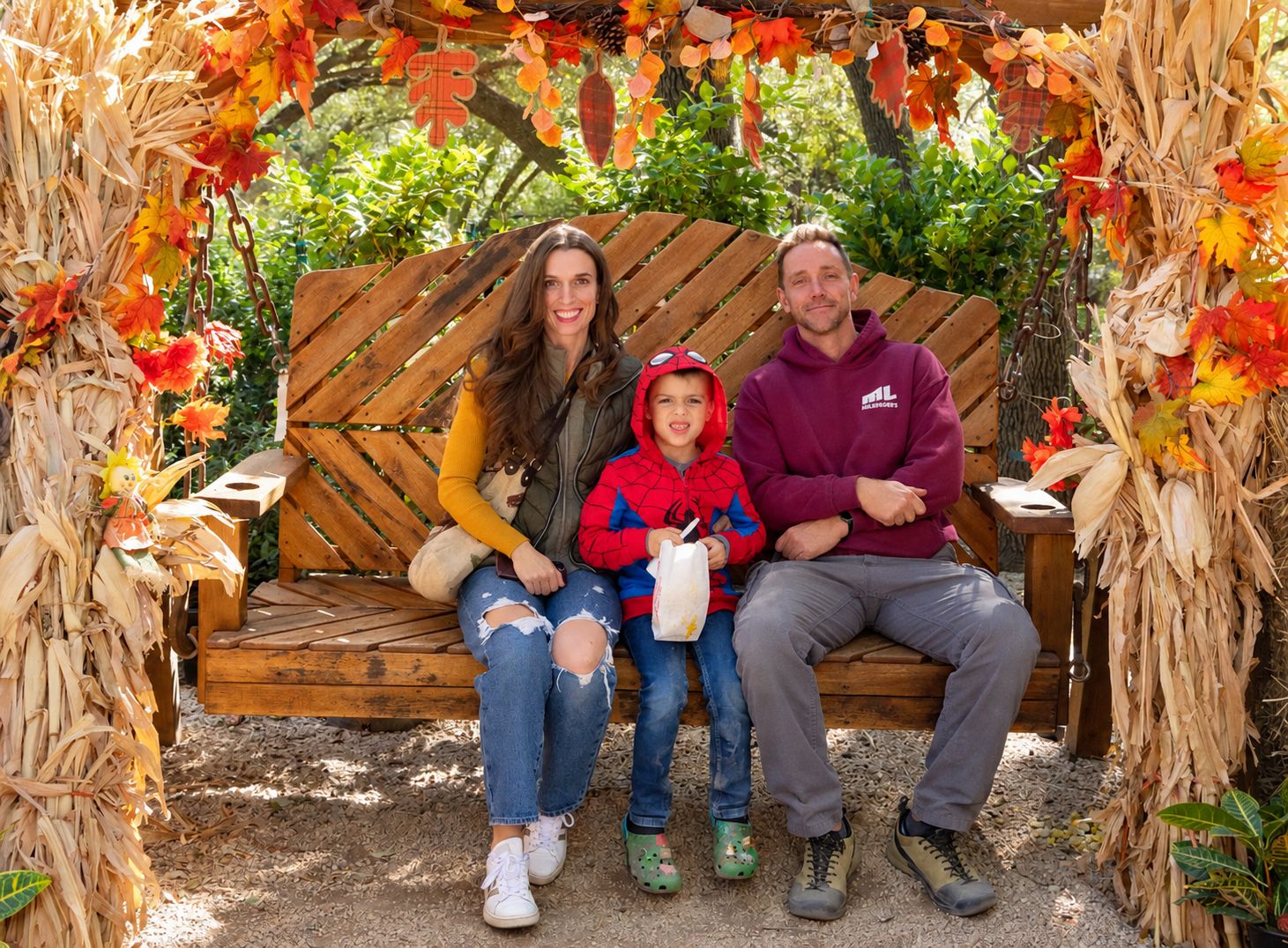 Smiling family of three sits on a wooden bench under a rustic autumn arch decorated with corn stalks and colorful fall leaves.