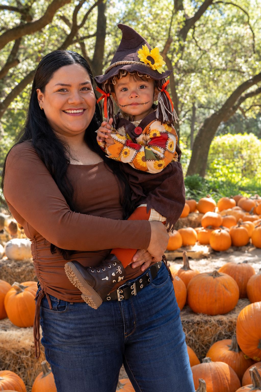 Smiling woman holding a toddler dressed as a scarecrow in a pumpkin patch with orange pumpkins, hay bales, and trees.