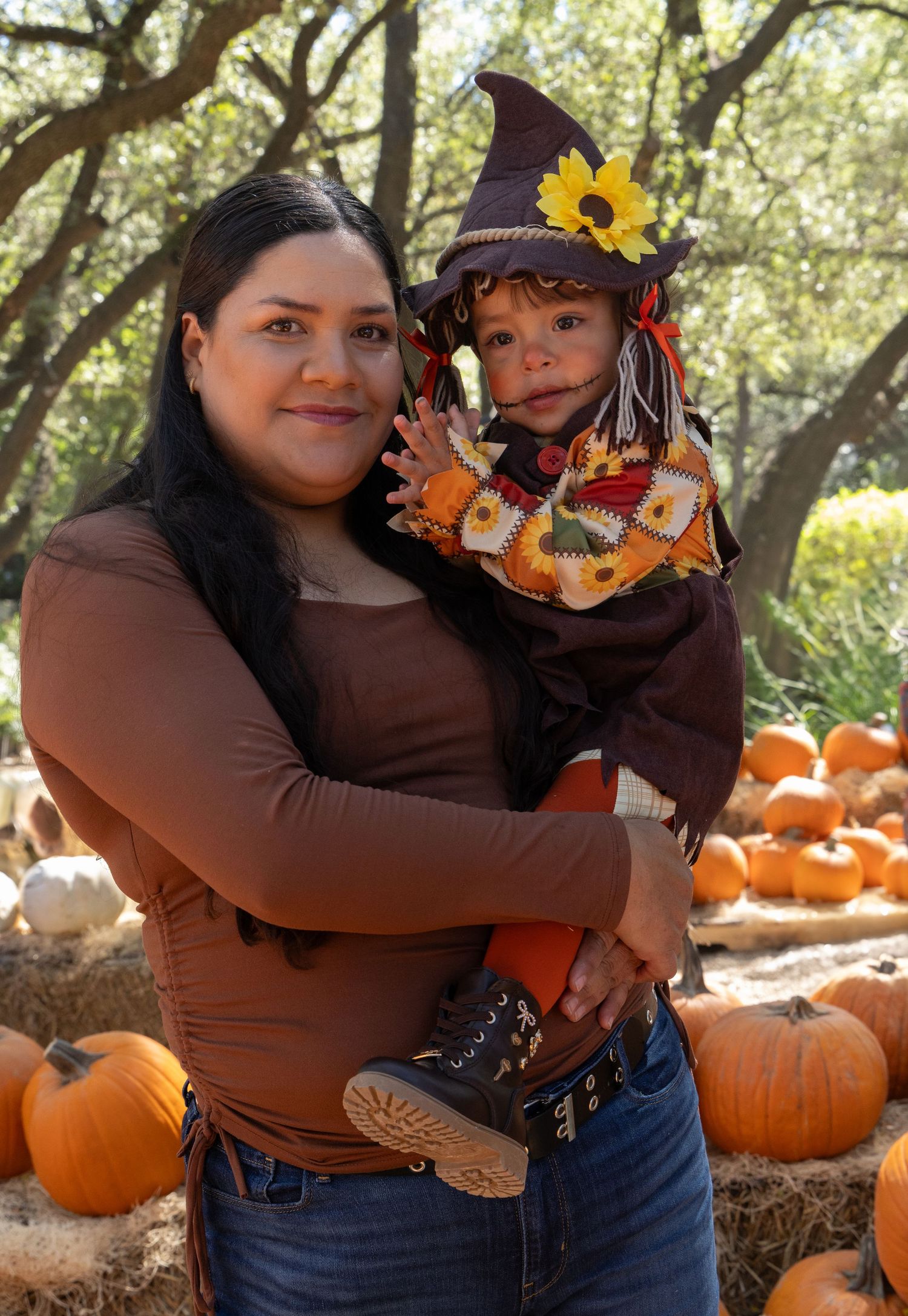 Smiling woman in a brown top holds a toddler dressed as a scarecrow with a witch hat near hay bales and pumpkins at a fall pumpkin patch.