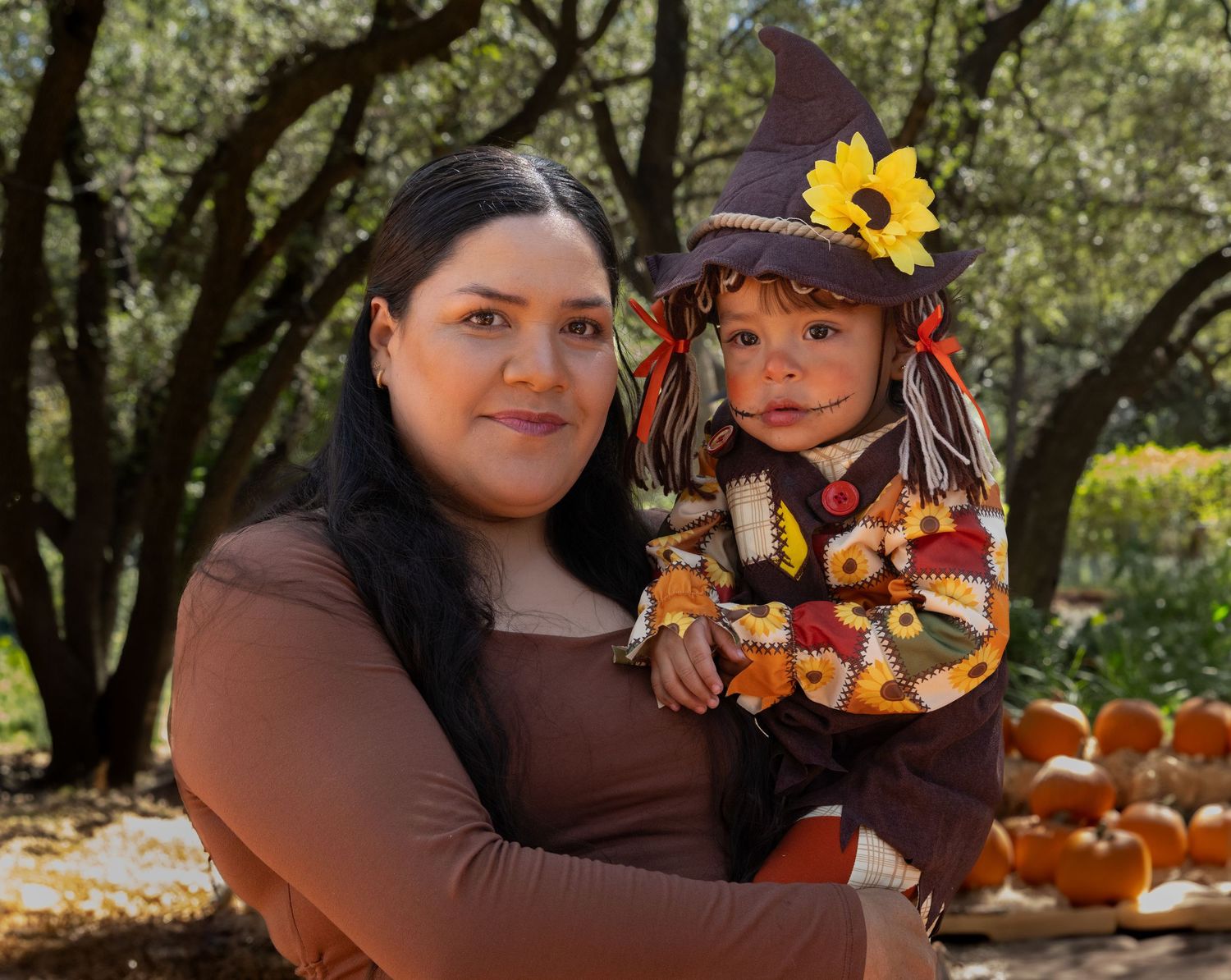 Smiling woman holds a toddler in a scarecrow costume with a witch hat and sunflower beside a pumpkin patch under shady trees.