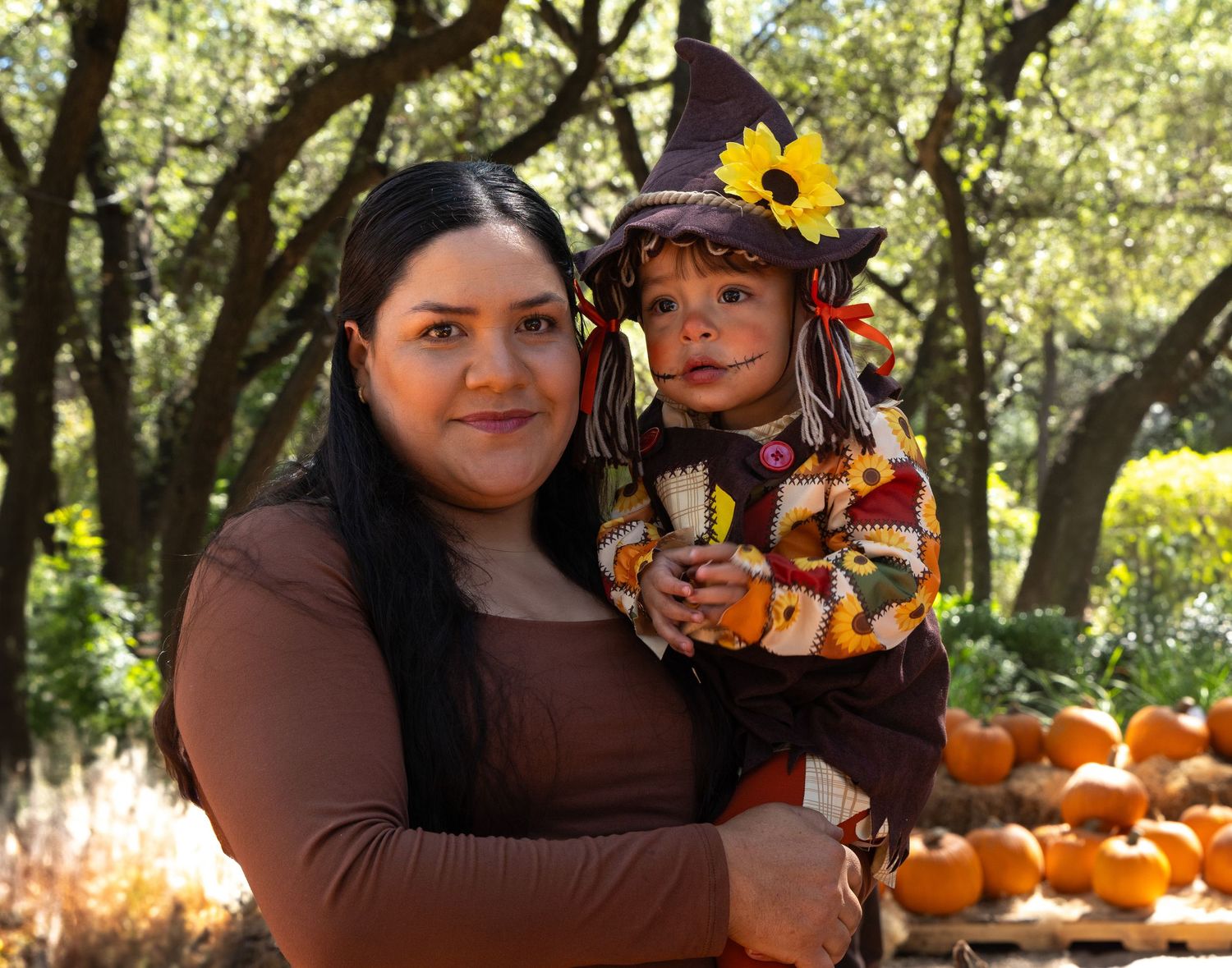 Smiling woman with long dark hair holds a toddler in a scarecrow costume with a sunflower hat beside a pumpkin patch under autumn trees.