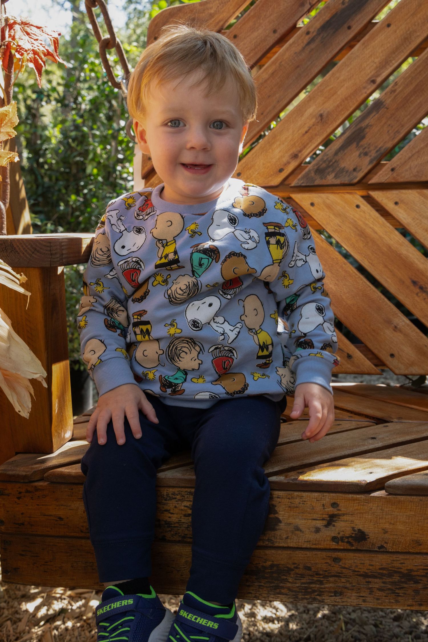 Smiling toddler in a blue Peanuts Snoopy and Charlie Brown sweatshirt, navy pants and Skechers sneakers sitting on a rustic wooden swing.