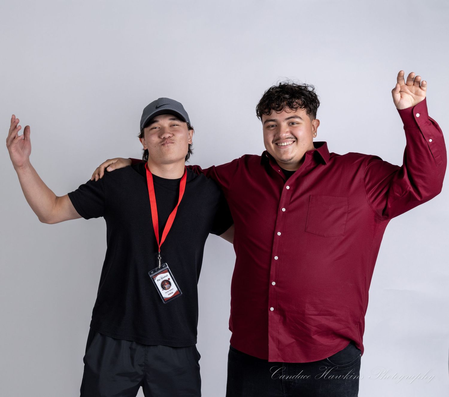 Two smiling young men pose arm in arm against a plain white backdrop, one in a black cap and lanyard, the other in a maroon button-up shirt.