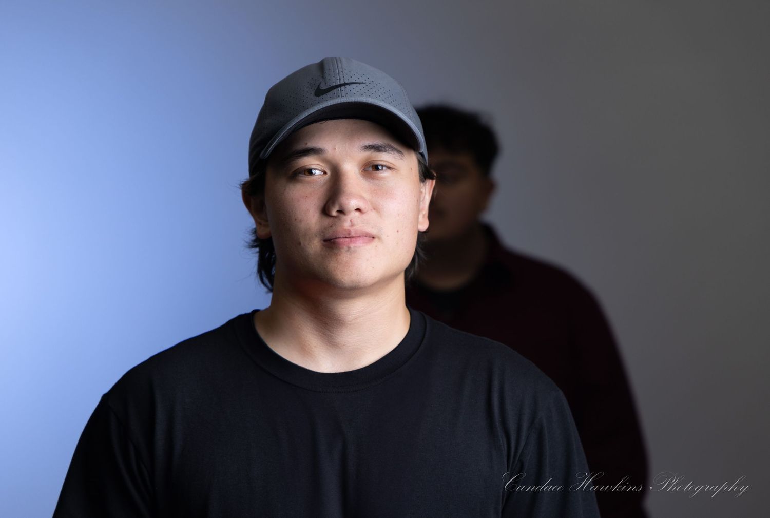 A young man in a gray cap and black t-shirt poses for a studio portrait against a blue background with a person behind him.