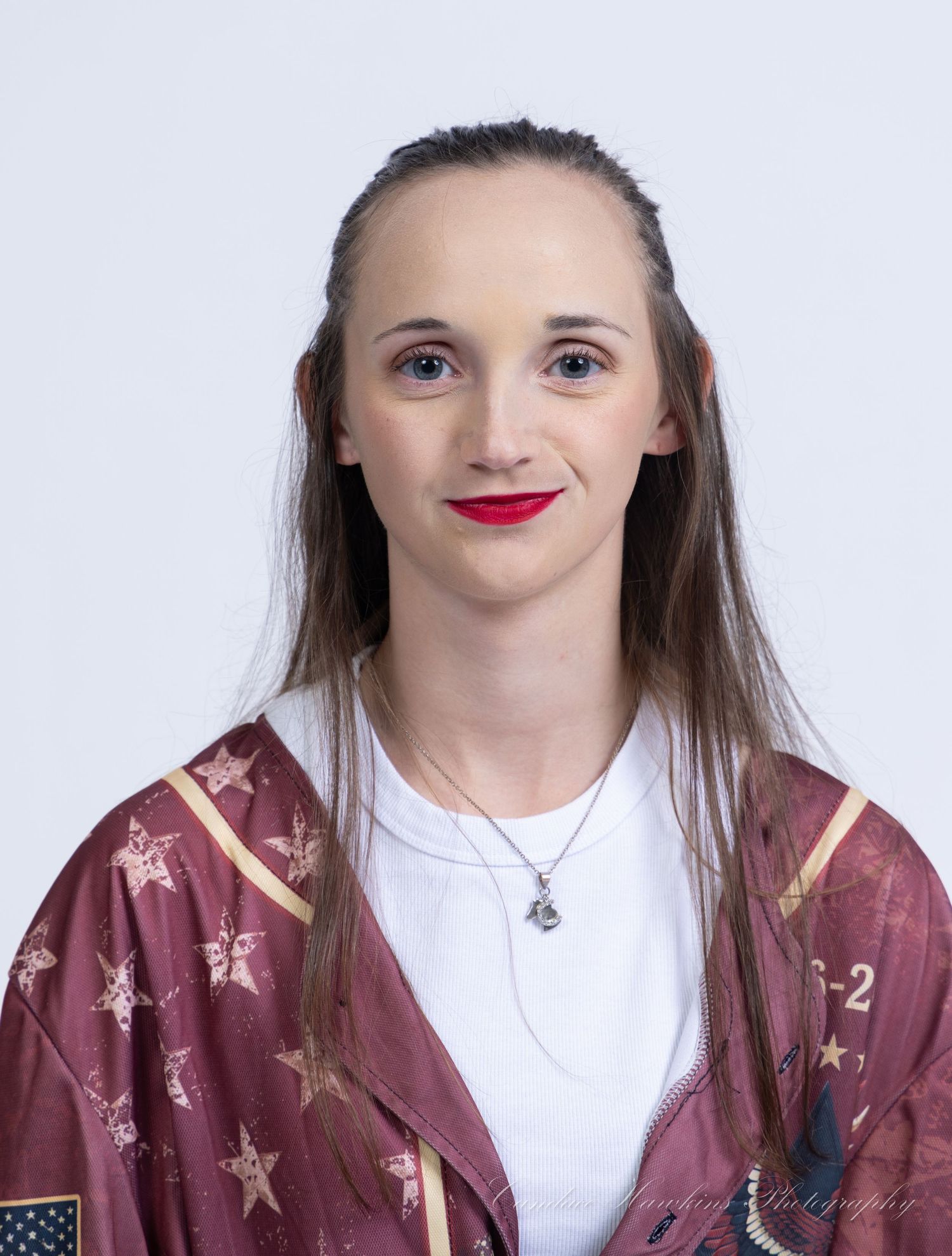 A studio headshot of a young woman with long brown hair and red lipstick wearing a maroon star-patterned jersey and necklace.