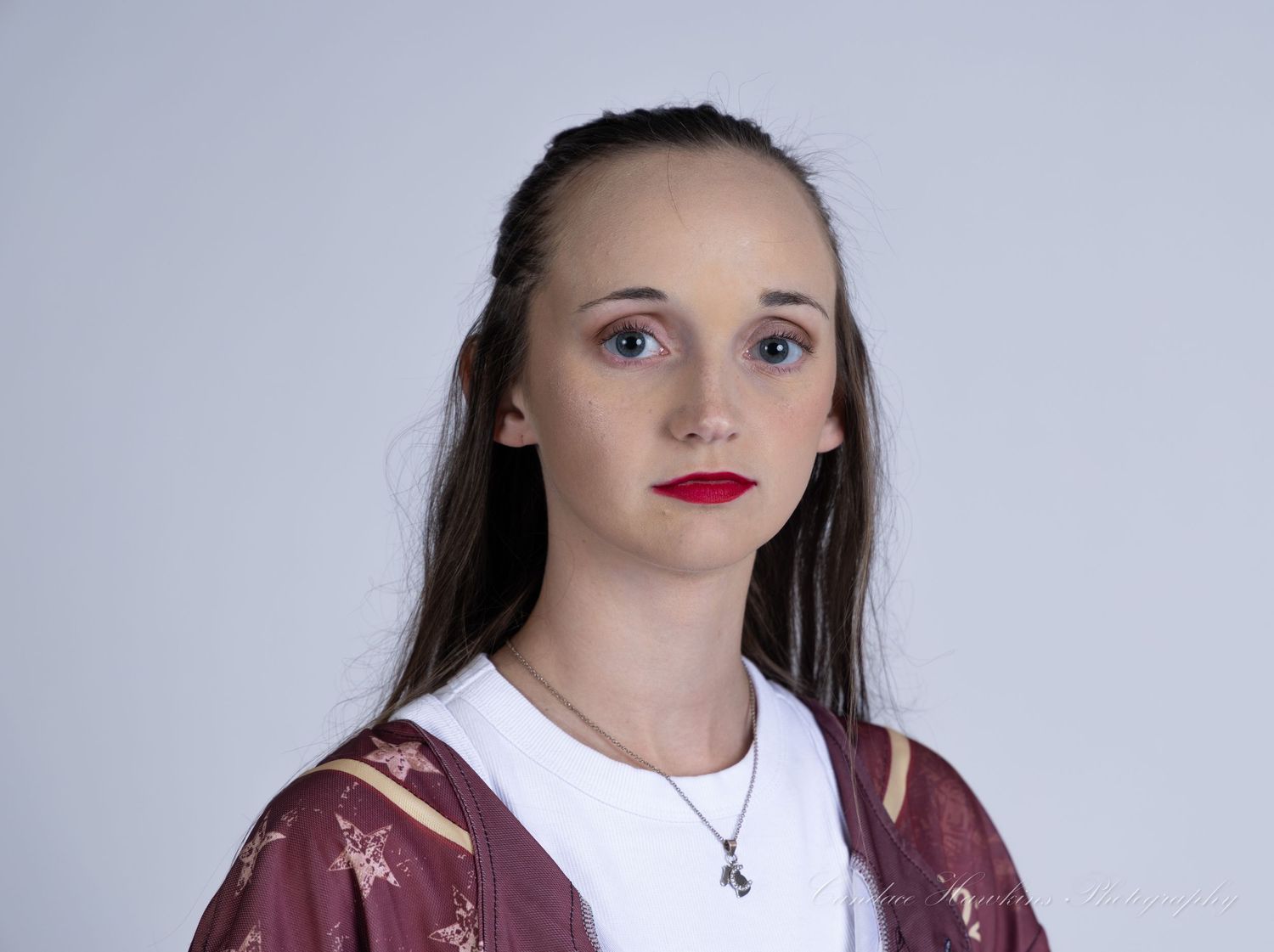 Young woman with long dark hair and red lipstick wears a white top, burgundy patterned jacket and a pendant necklace against a plain gray ba