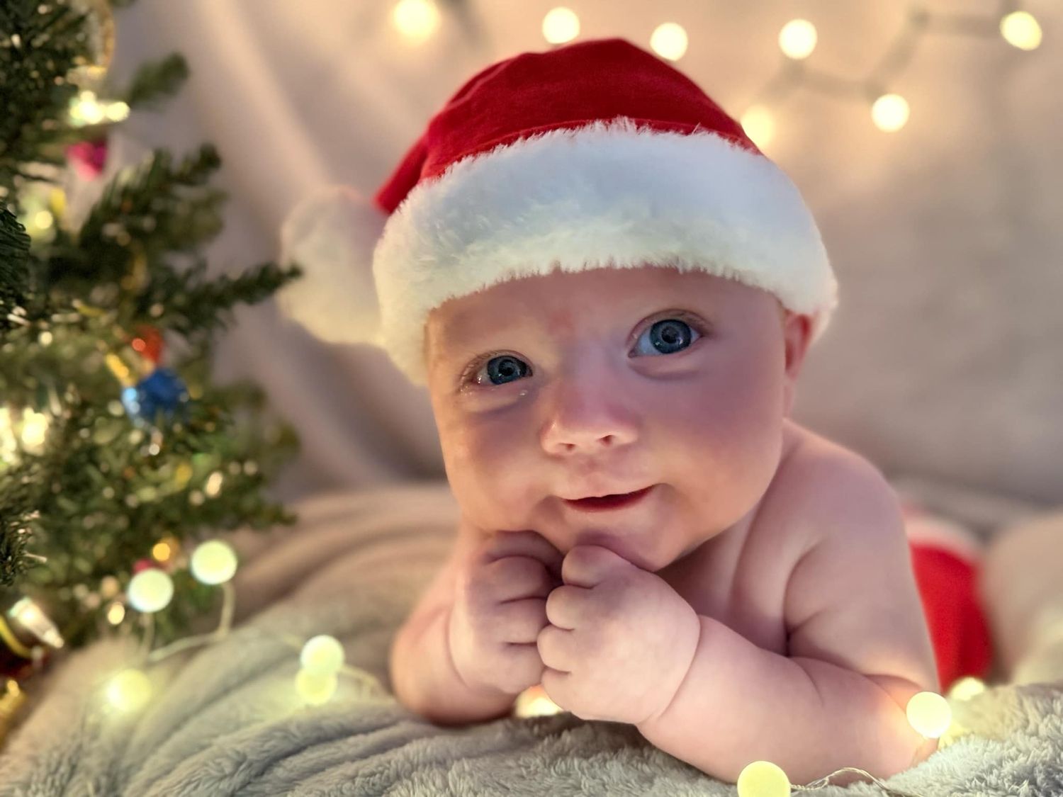 Baby with bright blue eyes wearing a Santa hat rests chin on tiny fists beside a Christmas tree and warm twinkling lights.