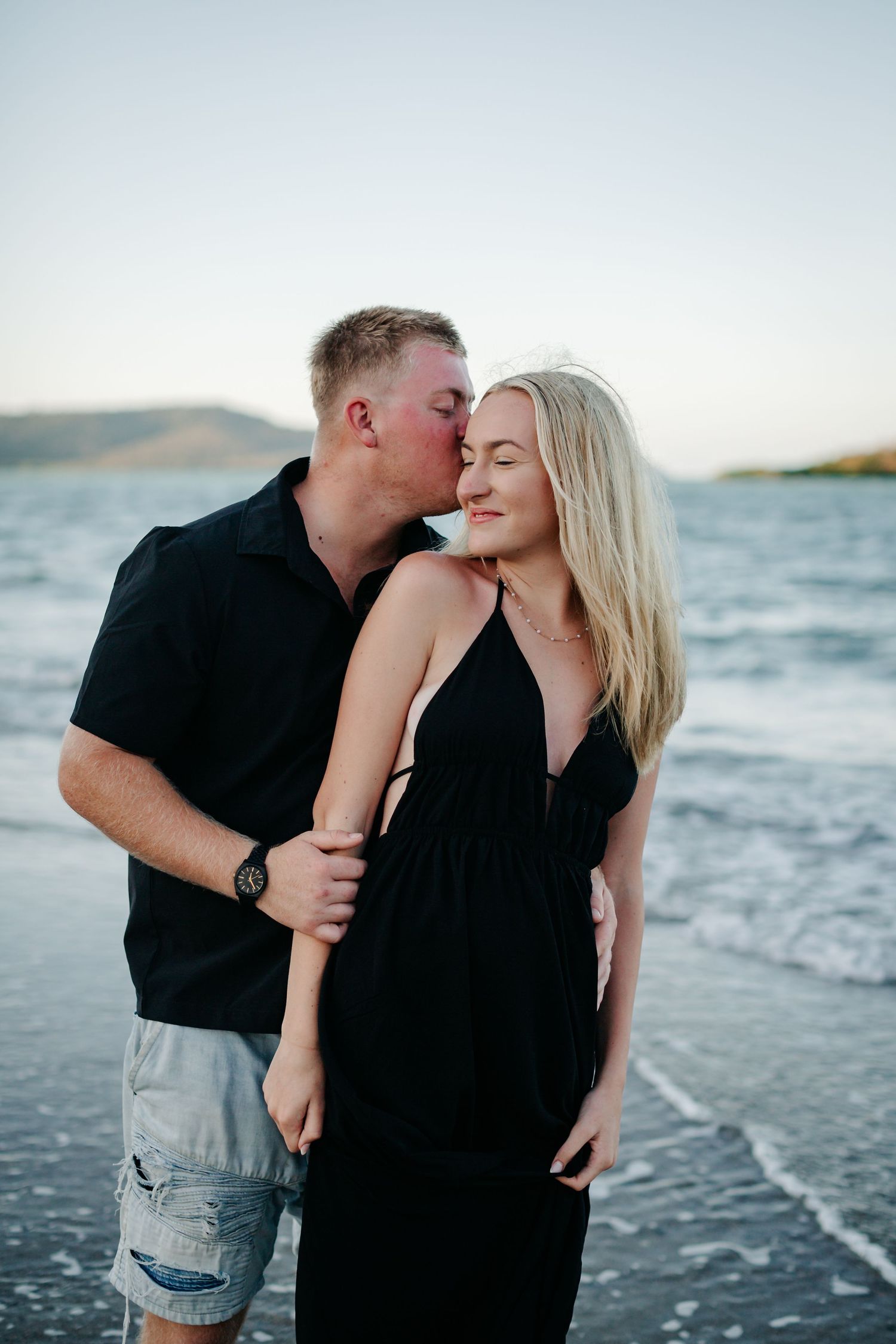 A couple embraces on a beach at sunset, man kissing woman's cheek, both wearing black outfits with ocean waves behind them.