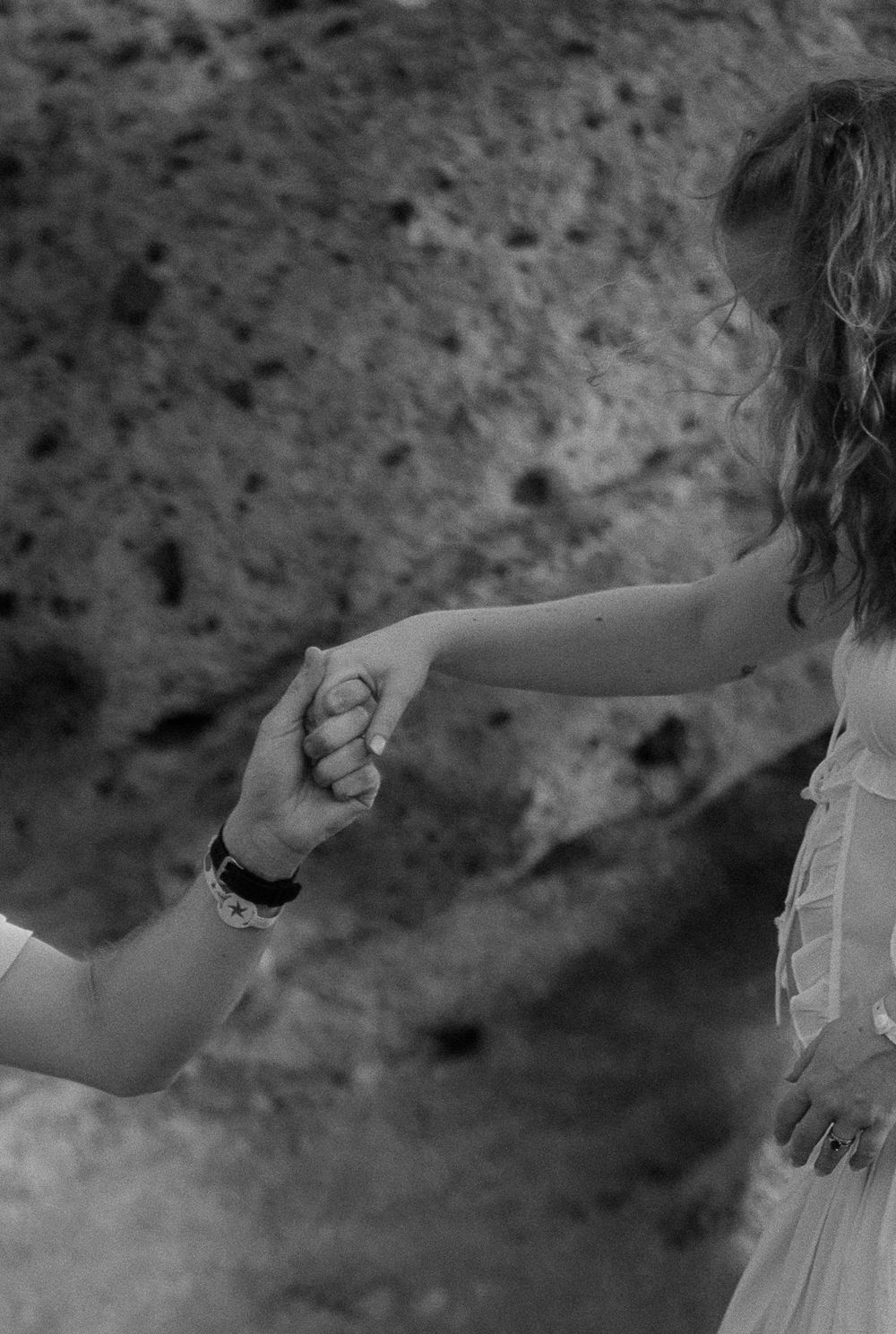Black and white photo of a couple holding hands tenderly near a rocky background.