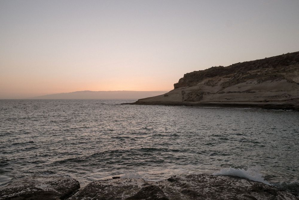 Serene coastal sunset with rocky cliffs meeting calm sea waters under a pastel sky.