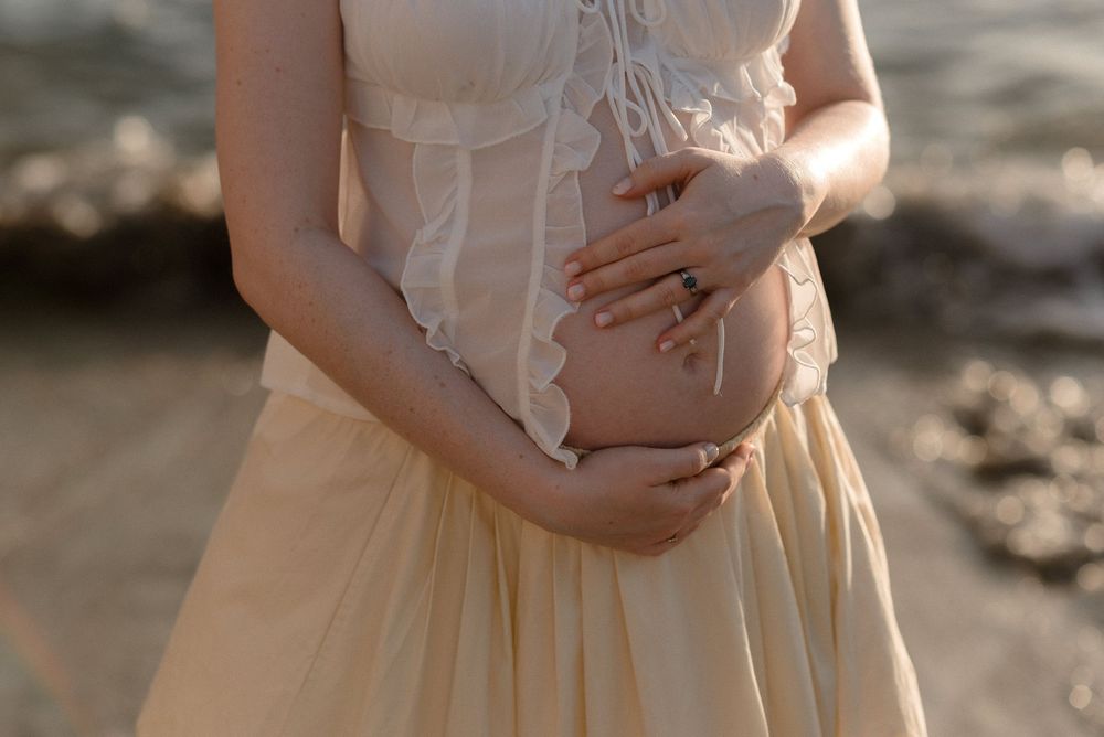 Pregnant woman in beige dress cradling baby bump near ocean shore in soft golden light.