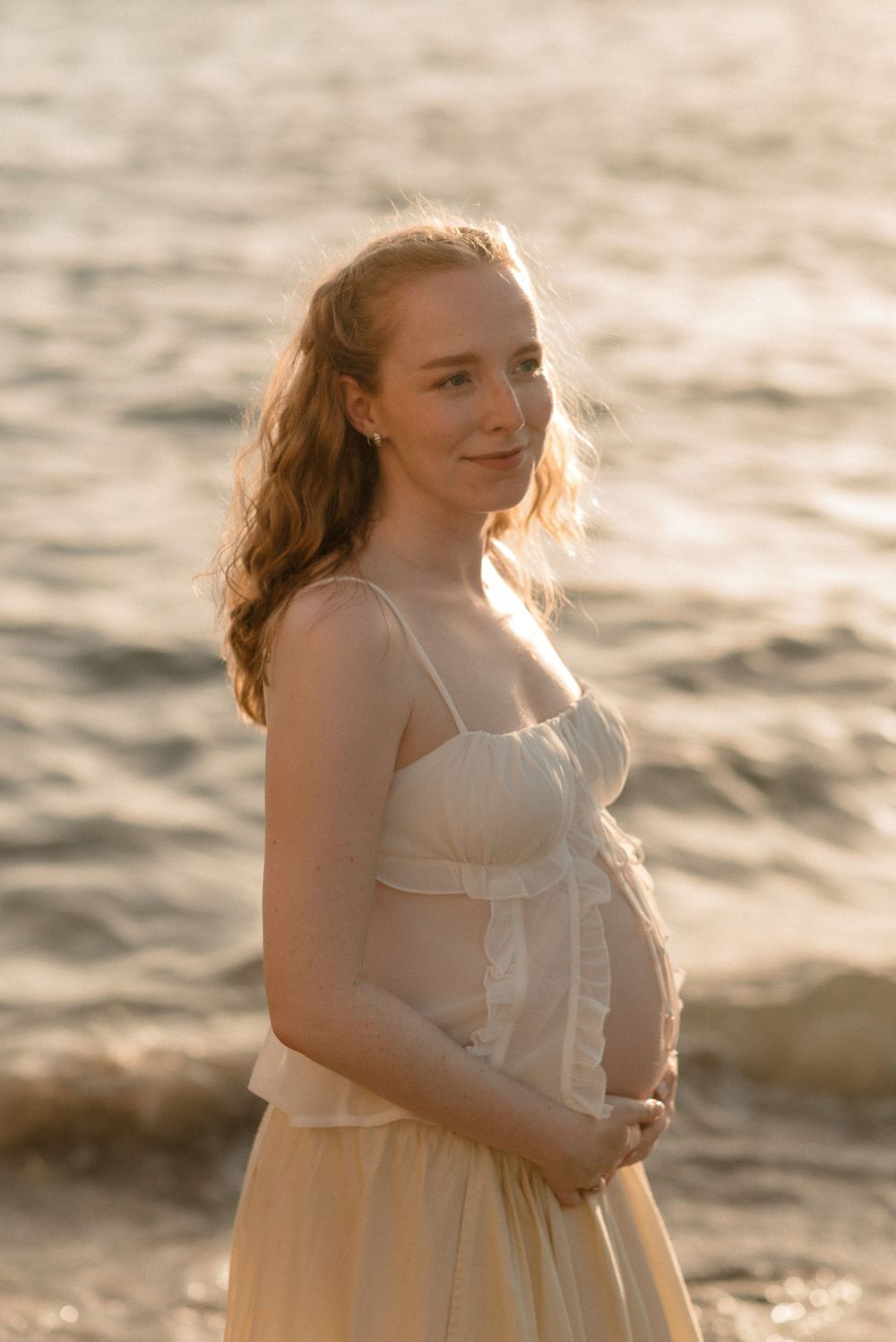 Pregnant woman in white dress standing on beach at golden hour, smiling softly with hands on belly.