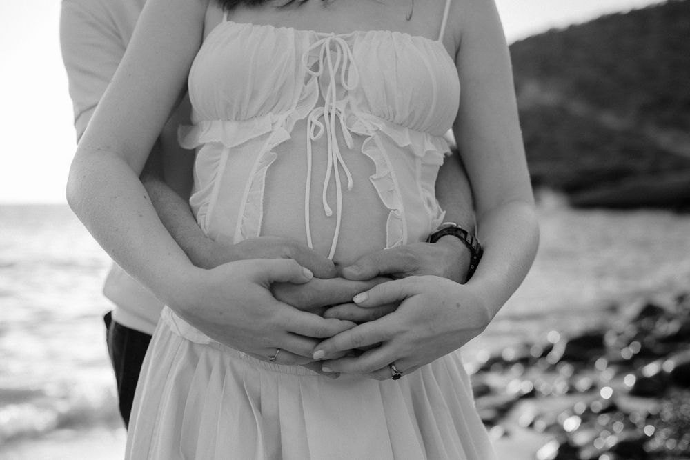 Black and white photo of a pregnant woman in a flowy dress with her partner cradling her baby bump on a rocky beach.
