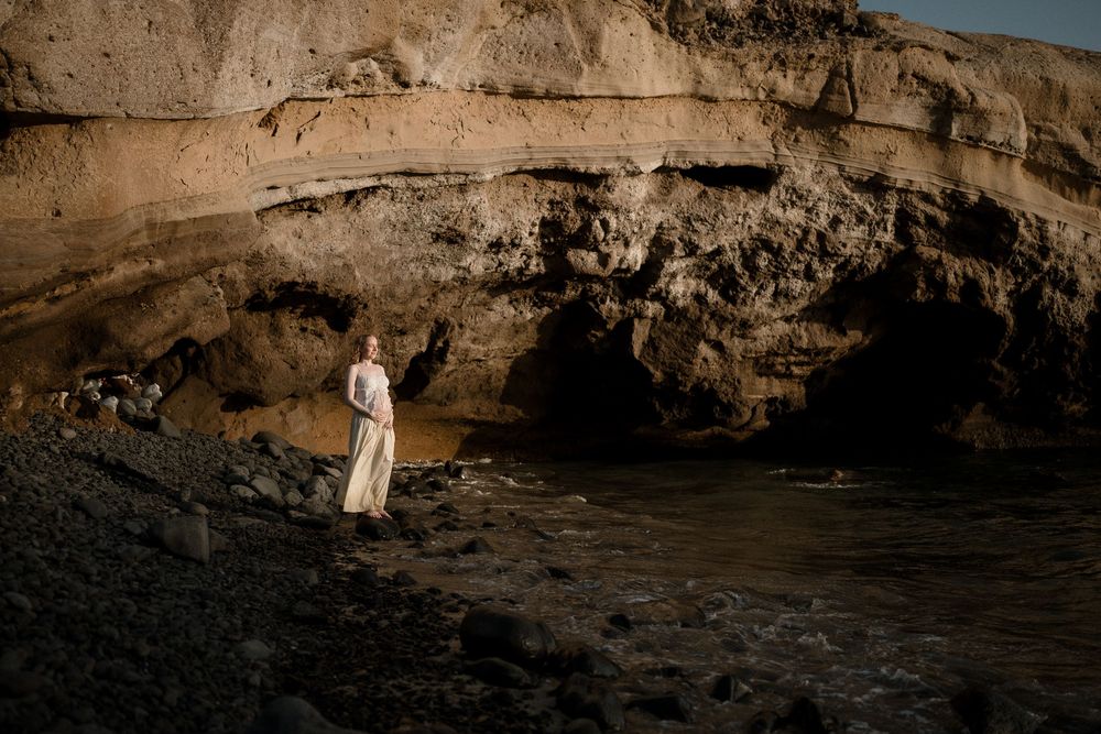 Woman in flowing dress stands by rocky coastal cave at golden hour.