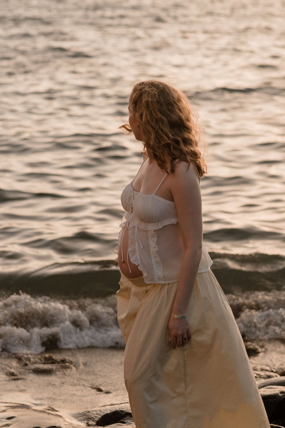 Pregnant woman in white crop top and beige skirt stands by ocean waves at golden hour sunset.
