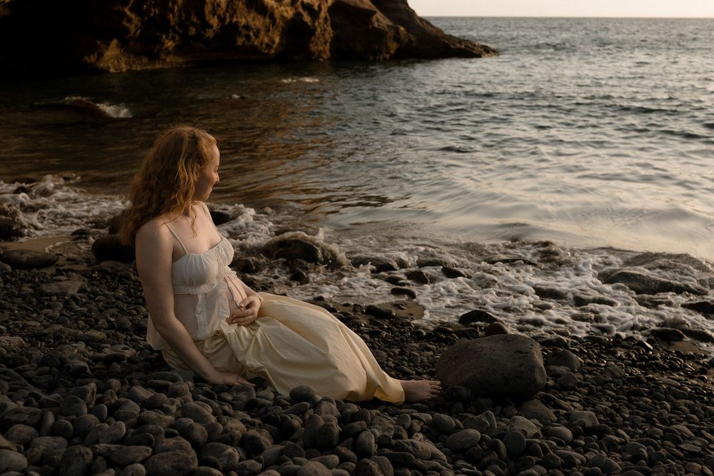 Woman in white dress sits on rocky beach at sunset by the ocean.