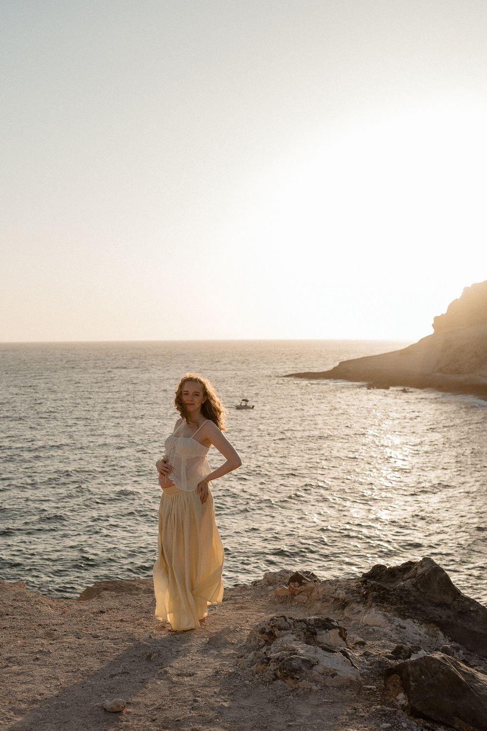 Woman in flowing cream dress standing on rocky coastal cliff at golden sunset with ocean and rocky headland behind her.