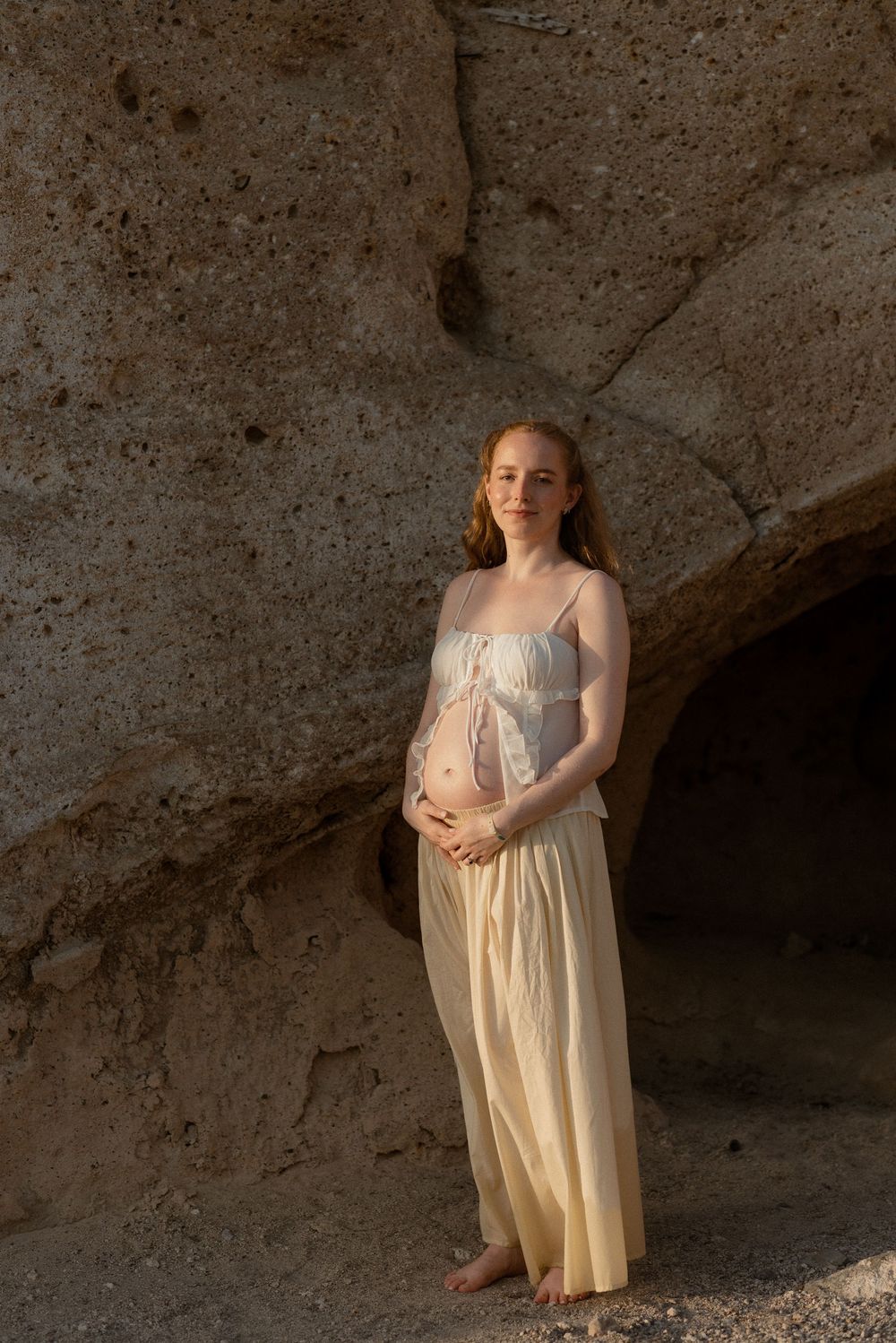 Pregnant woman in yellow skirt and white top stands barefoot against rocky cave wall in warm light.