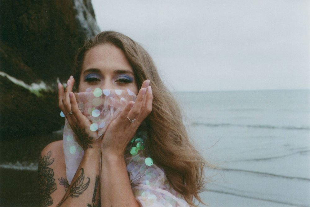 Woman with wavy hair and arm tattoos holds sparkling sequined fabric to her face beside a rocky cliff and calm gray ocean.