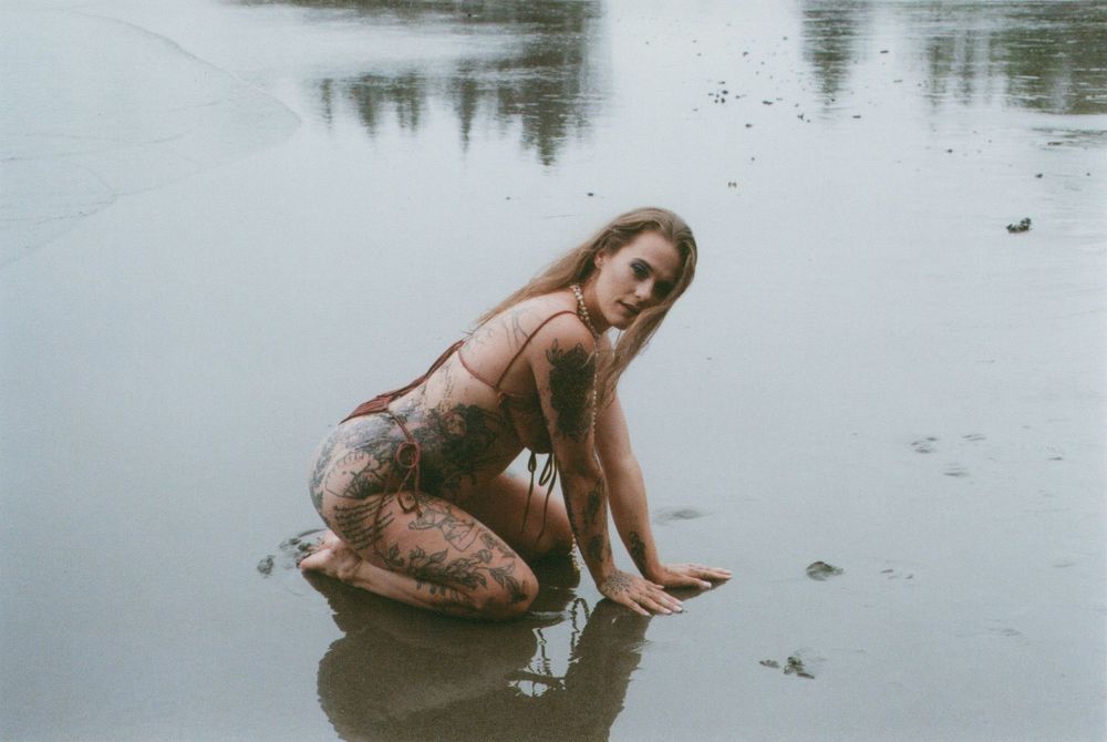 Woman with long blonde hair and arm tattoos kneeling in shallow water on a wet, reflective beach, touching the surface with one hand.
