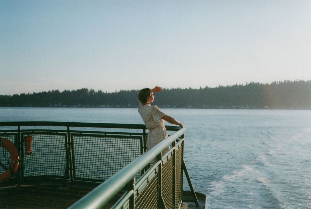 Woman in a light dress stands at the railing of a ferry deck, gazing over calm water toward a tree-lined shore at sunset.