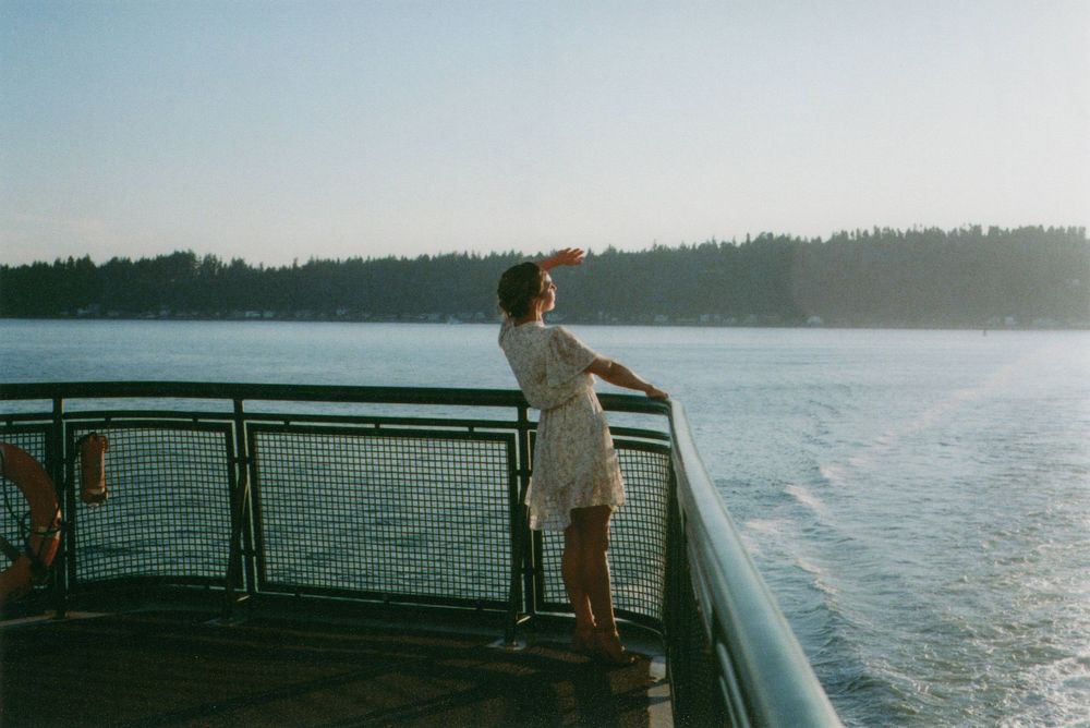Woman in a white dress stands at the railing of a ferry deck, shielding her eyes while gazing over calm water toward a tree-lined shore.