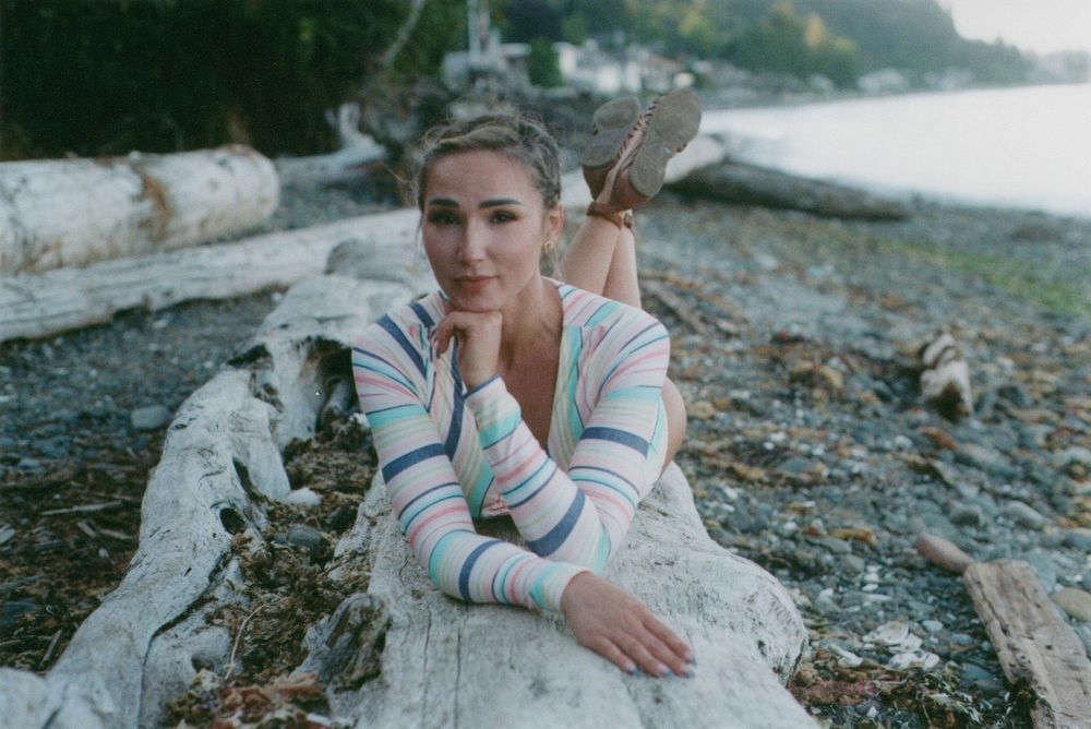Woman in a striped sweater lies on driftwood along a rocky beach, resting her chin on her hand with calm water and trees behind her.