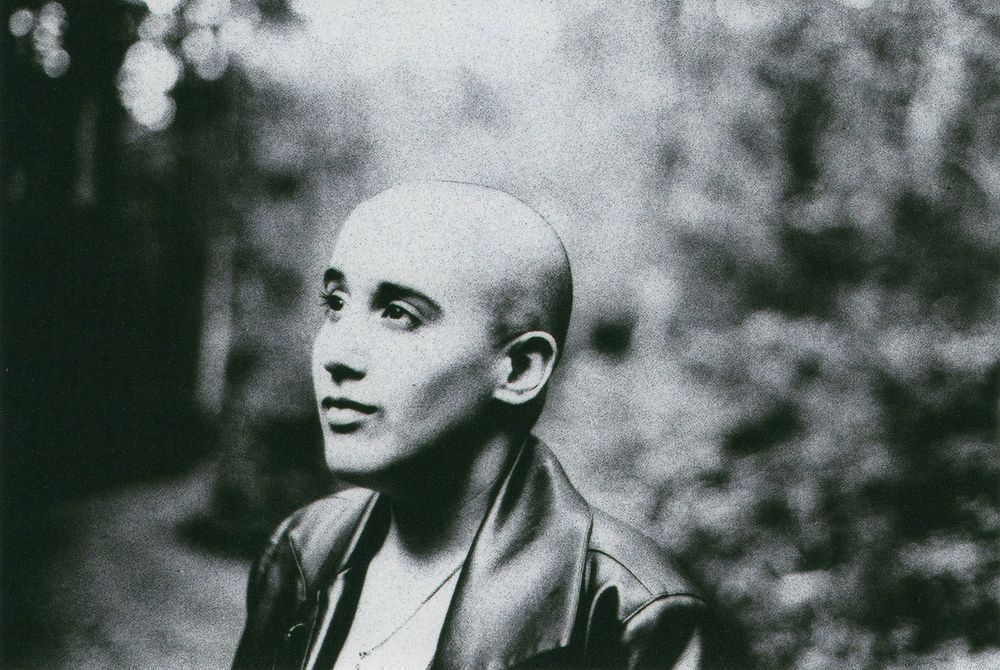 Black and white portrait of a bald person in a leather jacket gazing upward in a softly blurred, tree-filled outdoor setting.
