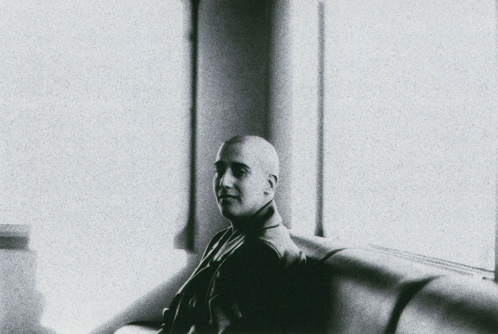 Black and white photo of a bald man in a dark jacket sitting on a curved bench beside large windows, glancing toward the camera.