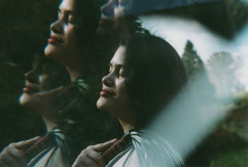 Double exposure portrait of a woman with closed eyes and dark hair, face tilted upward in soft light, hands raised near her chest.