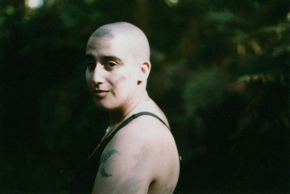 Person with a shaved head and a shoulder tattoo glancing sideways in a dark, leafy forest, wearing a dark tank top in soft natural light.