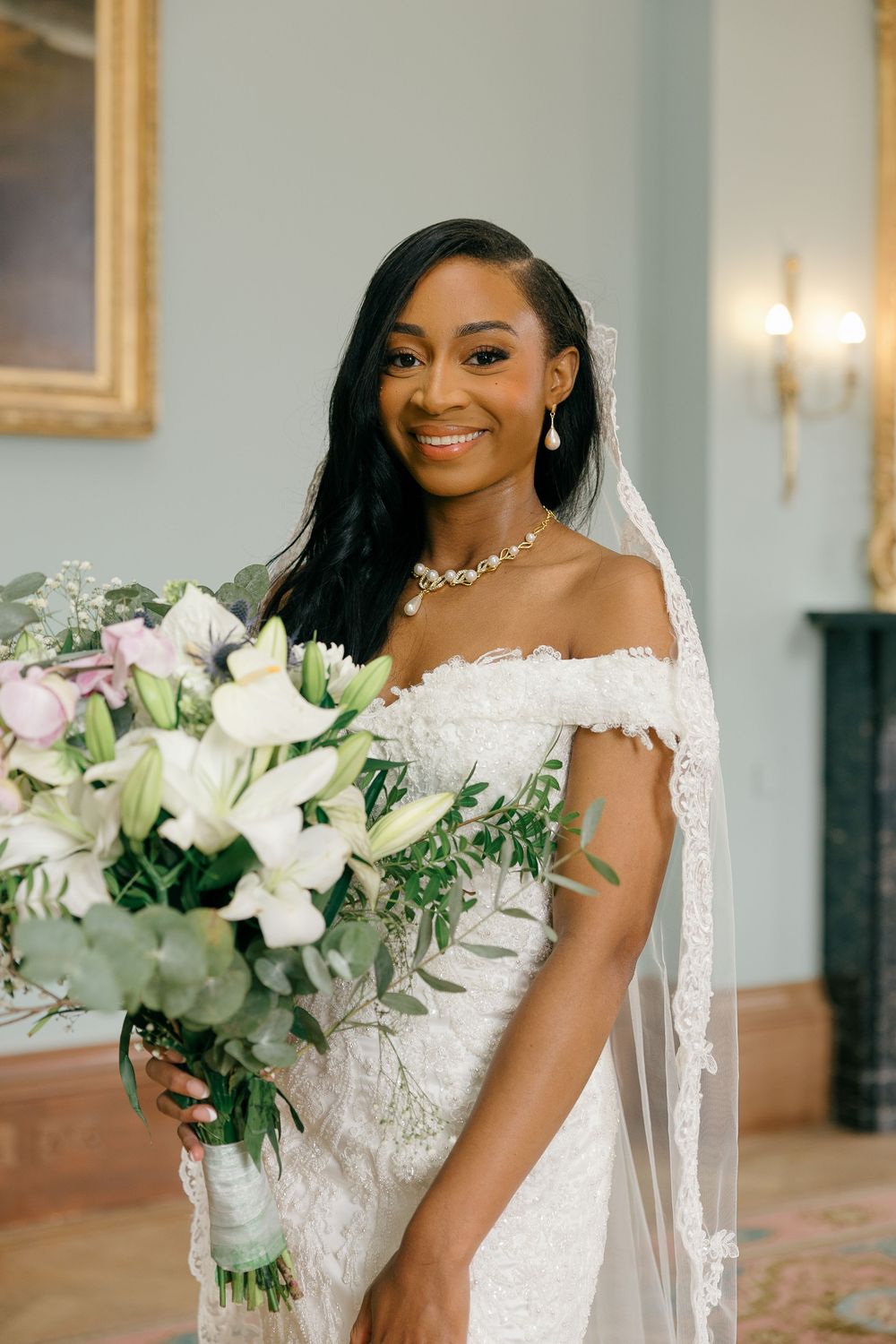 Smiling Black bride in off-shoulder lace gown holds white floral bouquet with veil in elegant blue room.
