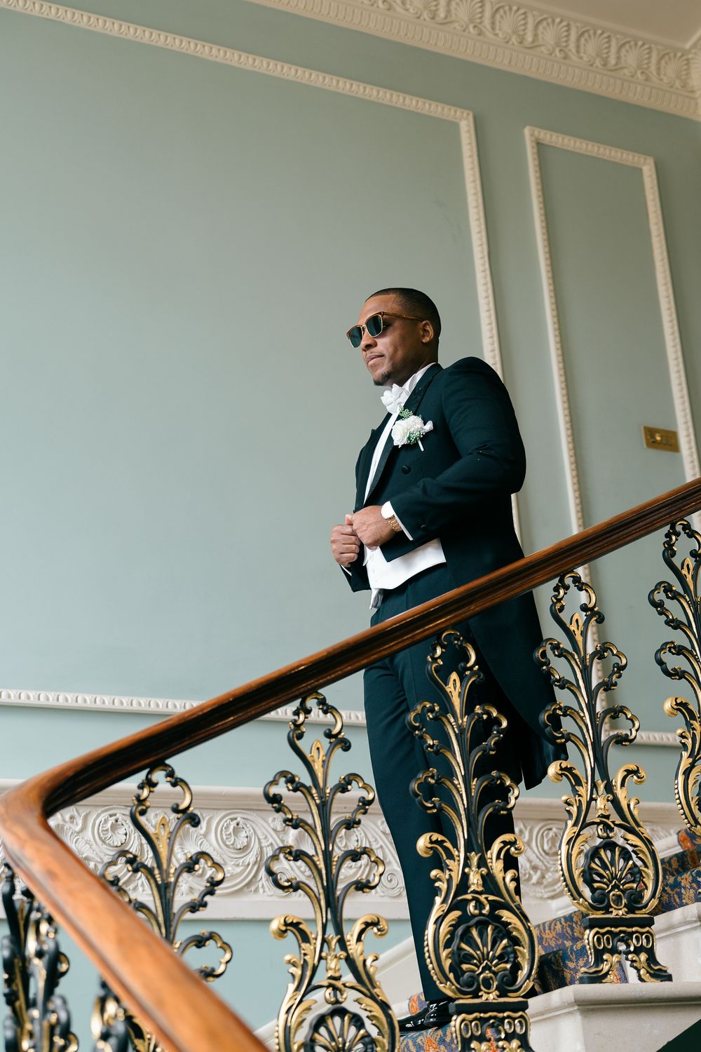Elegant groom in black tuxedo and sunglasses stands on ornate golden staircase in grand venue.