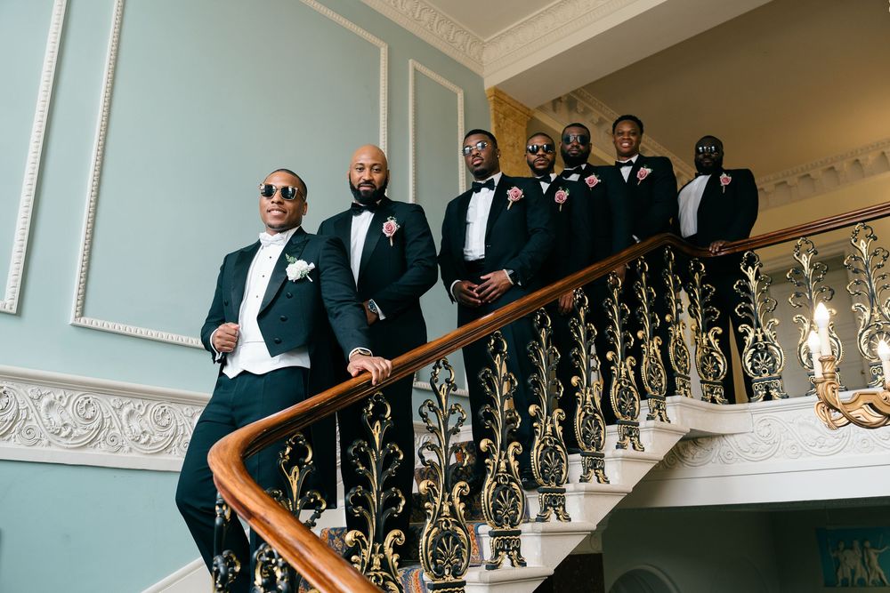 Groomsmen in black tuxedos pose on an elegant staircase with gold railings at a wedding venue.