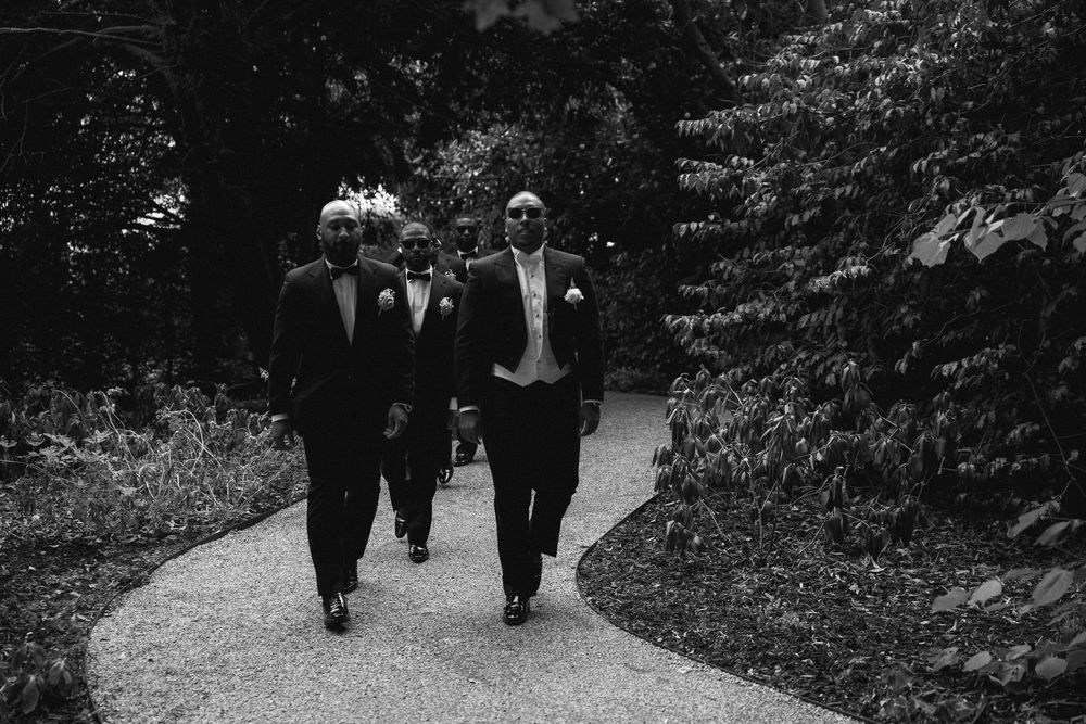 Black and white photo of groomsmen in tuxedos walking down a garden path at a wedding.