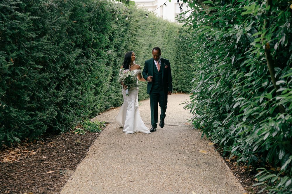 Bride and groom walk hand in hand along a lush garden path lined with tall green hedges.