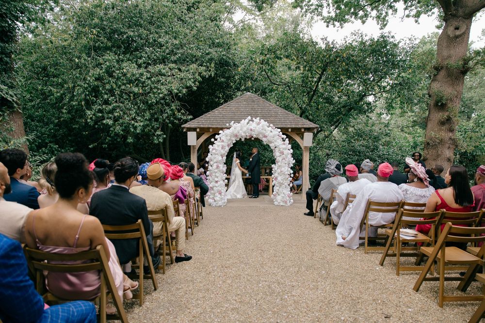 Outdoor wedding ceremony with floral arch and guests seated on wooden chairs in garden.