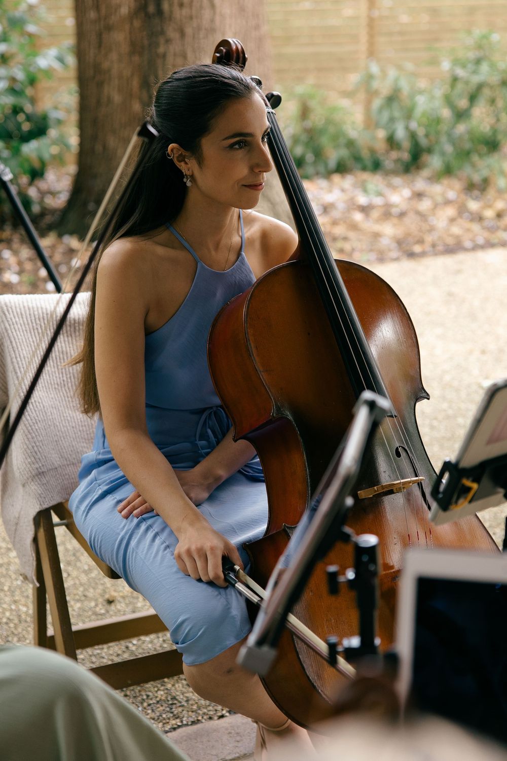 A woman in a blue outfit plays cello outdoors at a wedding reception.