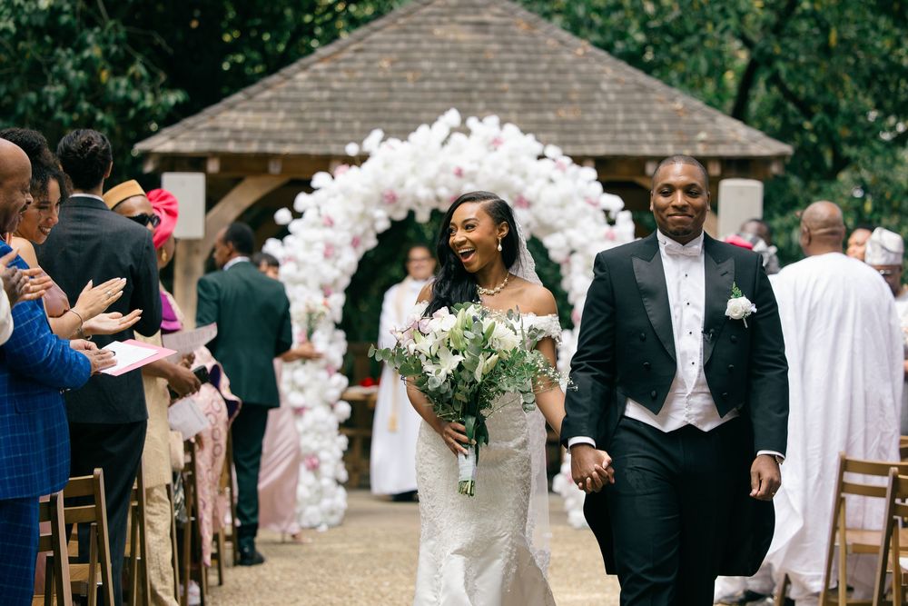 Bride and groom walk down aisle after ceremony under white floral arch in garden venue.