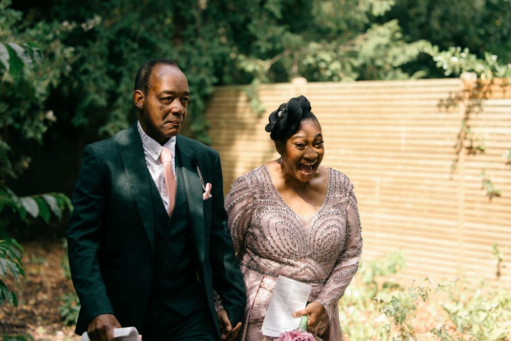 Elegant couple in formal attire walking outdoors, man in dark suit with pink tie, woman in beaded gown, at a wedding.